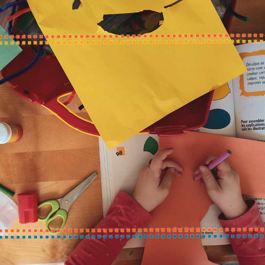 From a bird’s eye view, a pair of light skinned children’s hands hold a purple crayon. The hands are placed on top of a table filled with crafts material like construction paper and a craft book. The child appears to be drawing on cut orange paper. To the left of the child is a pair of scissors and a pencil sharpener. Rows of orange, yellow, and blue square dots serve as a border on the top and bottom of the image.
