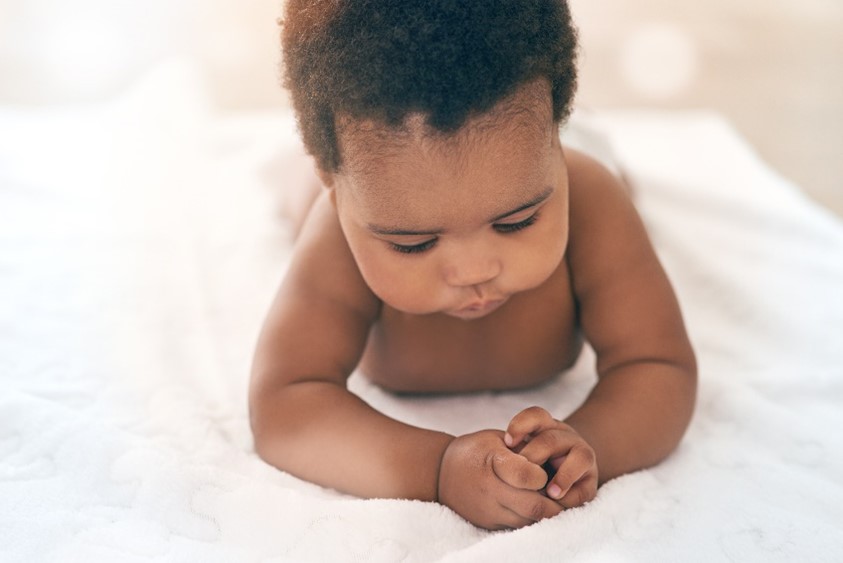 A baby laying facing forward on a white blanket holding their hands together.