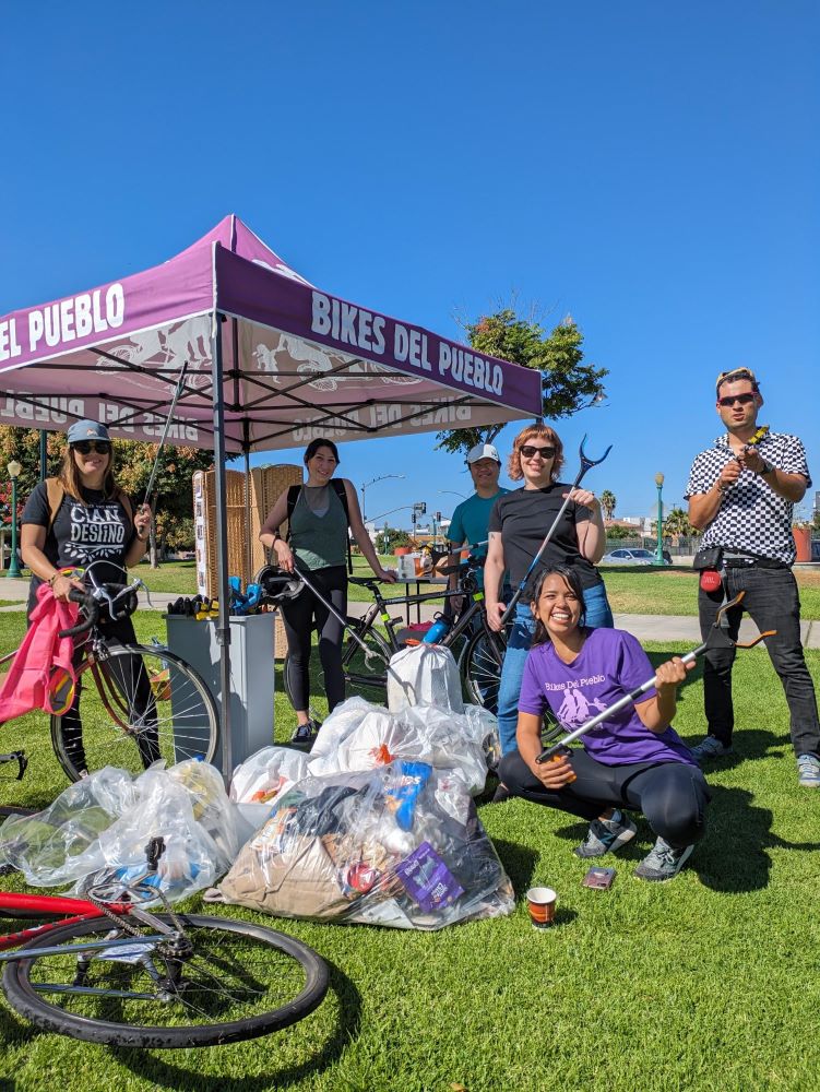 Six people next to a canopy that is labeled Bikes Del Pueblo. The people are posed next to trash bags.