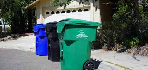 A blue trash bin, a black trash bin and a green trash bin next to a curb