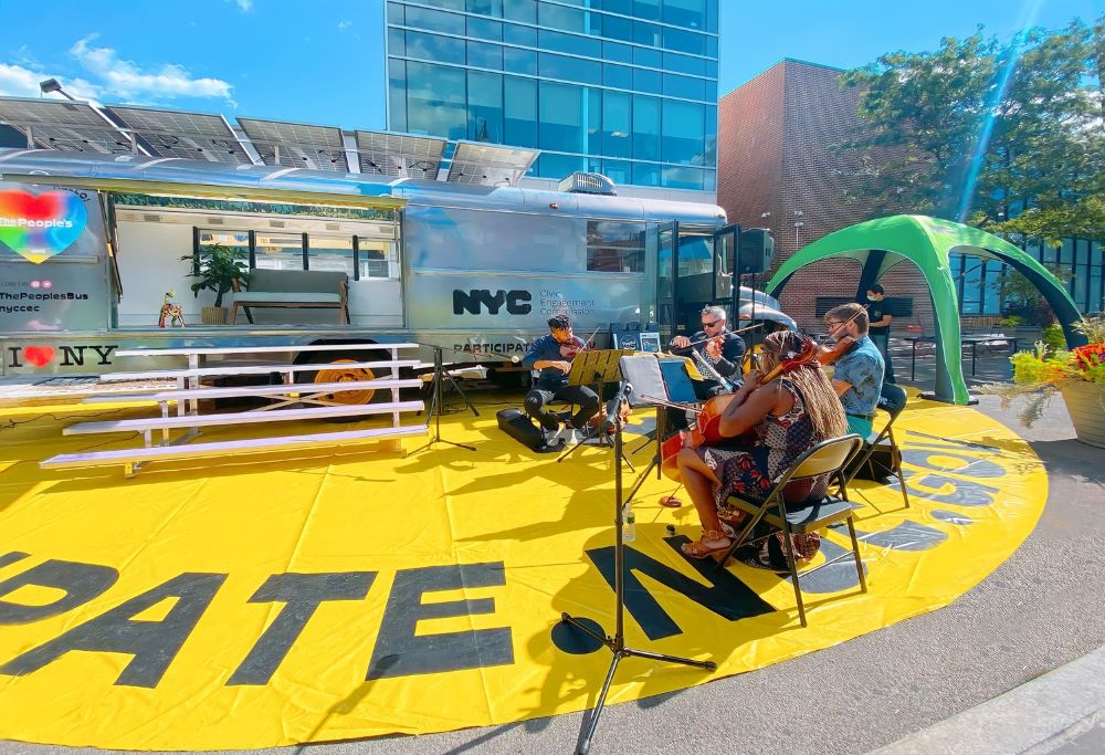 Four musicians play instruments in front of a New York City People's Bus. The bus is gray and displays a couch within the bus and stairs up to the bus.