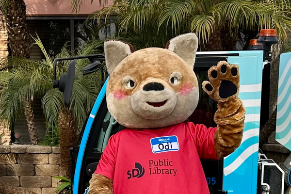 Library Mascot Odi the Coyote waves at the camera on an outdoor set