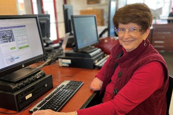 An older woman in glasses is seated at a computer workstation with Ancestry.com on the screen