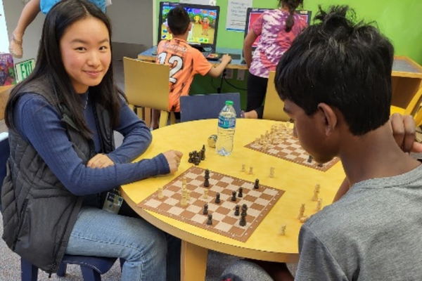 A teen playing chess against a young boy smiles at the camera while waiting for her turn