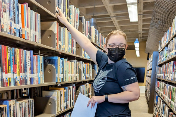 A masked volunteer holds a list in one hand while reaching for a book with the other