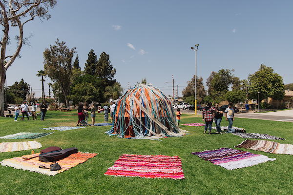 A colorful dome structure surrounded by colorful blankets spread out on grass.