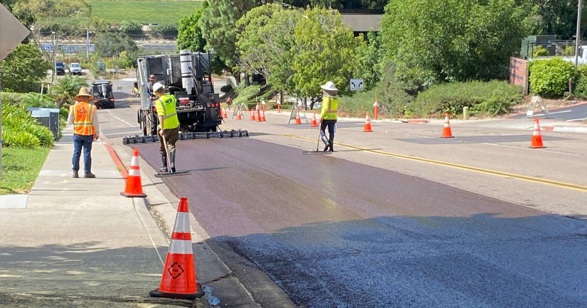 three workers slurry sealing a section of a street&#44; surrounded by trees