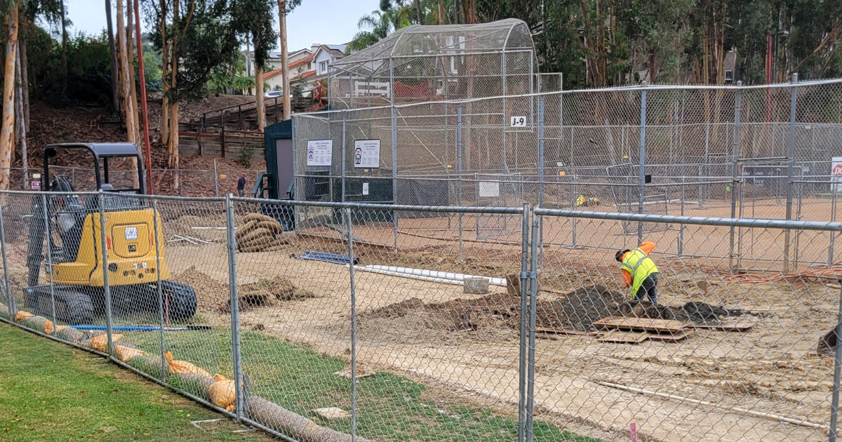 construction worker digging dirt surrounded by a chain fence and baseball field