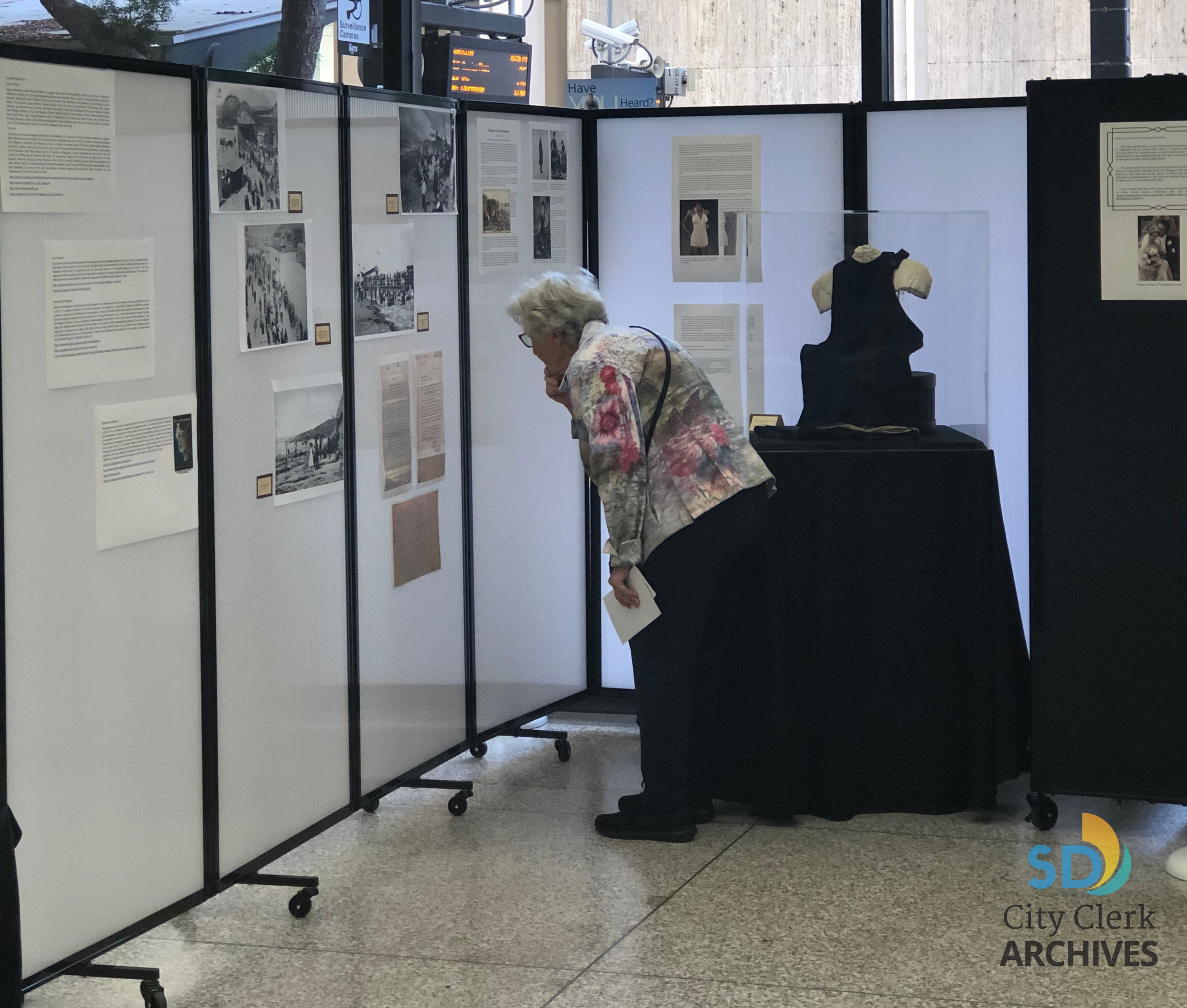 Elderly woman reading the articles posted on a white wall with a 1920s woman's blue wool bathing costume behind her.