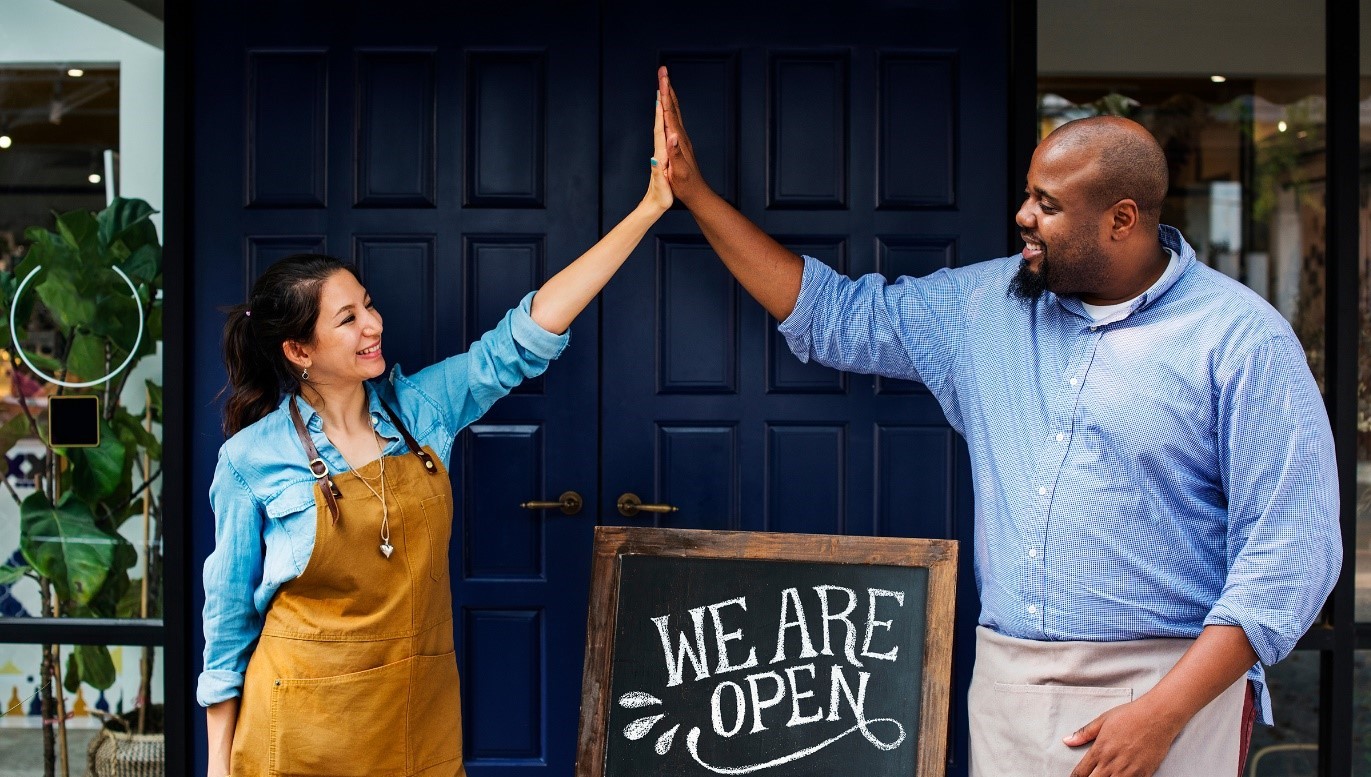 Two small business owners high-five each other in front of a "We are Open" sign.