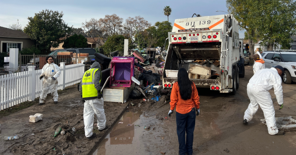 five people working to clear out trash in a trash truck on the street. 
