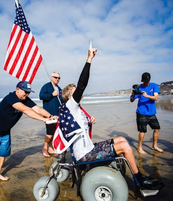 Photo of person being pushed in a manual beach wheelchair through wet sand near the ocean.