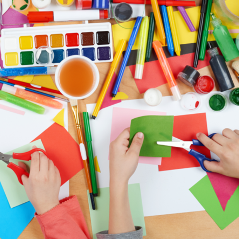 Two kids hands on top of a table with paper, scissors and other art supplies.