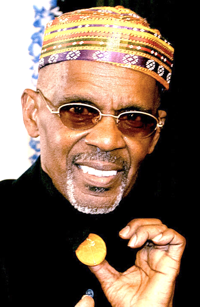 An image of an older black gentleman in tinted glasses with a white and gray goatee, a colorful hat, and fingers a golden pendant at his collar.