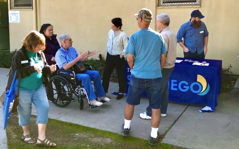 Artist Kate Clark at a table with people standing around her as she talks about her art project