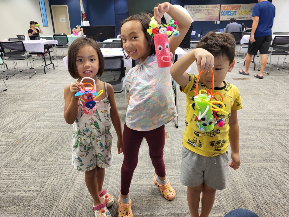 Three kids smiling holding up colorful crafts made with paper and pipe cleaners.