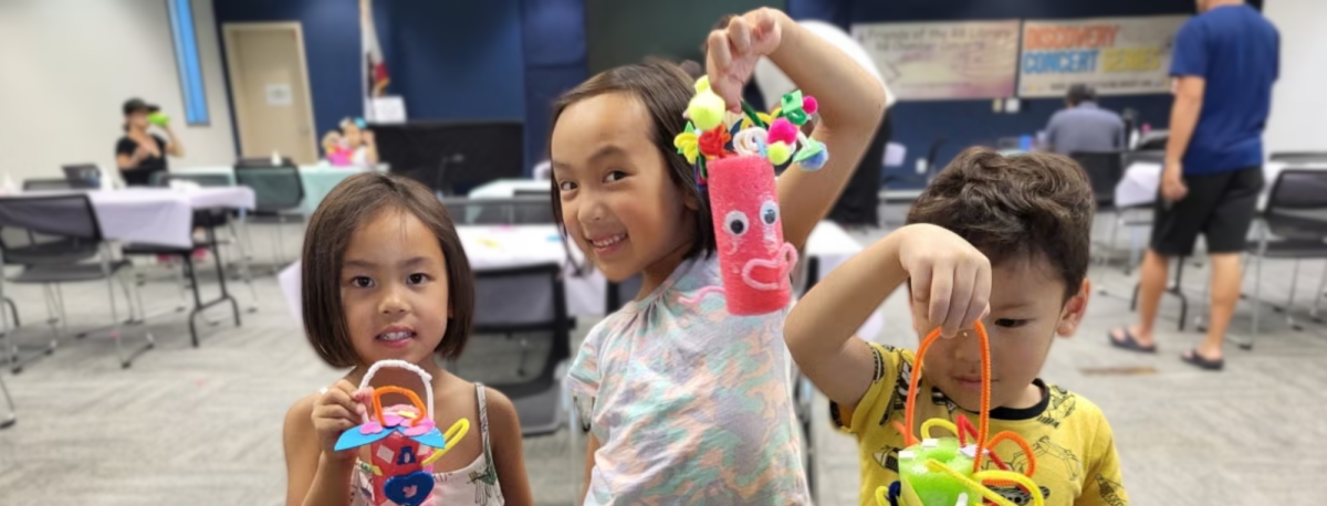 Three happy young children hold up colorful pool foam crafts at the library.