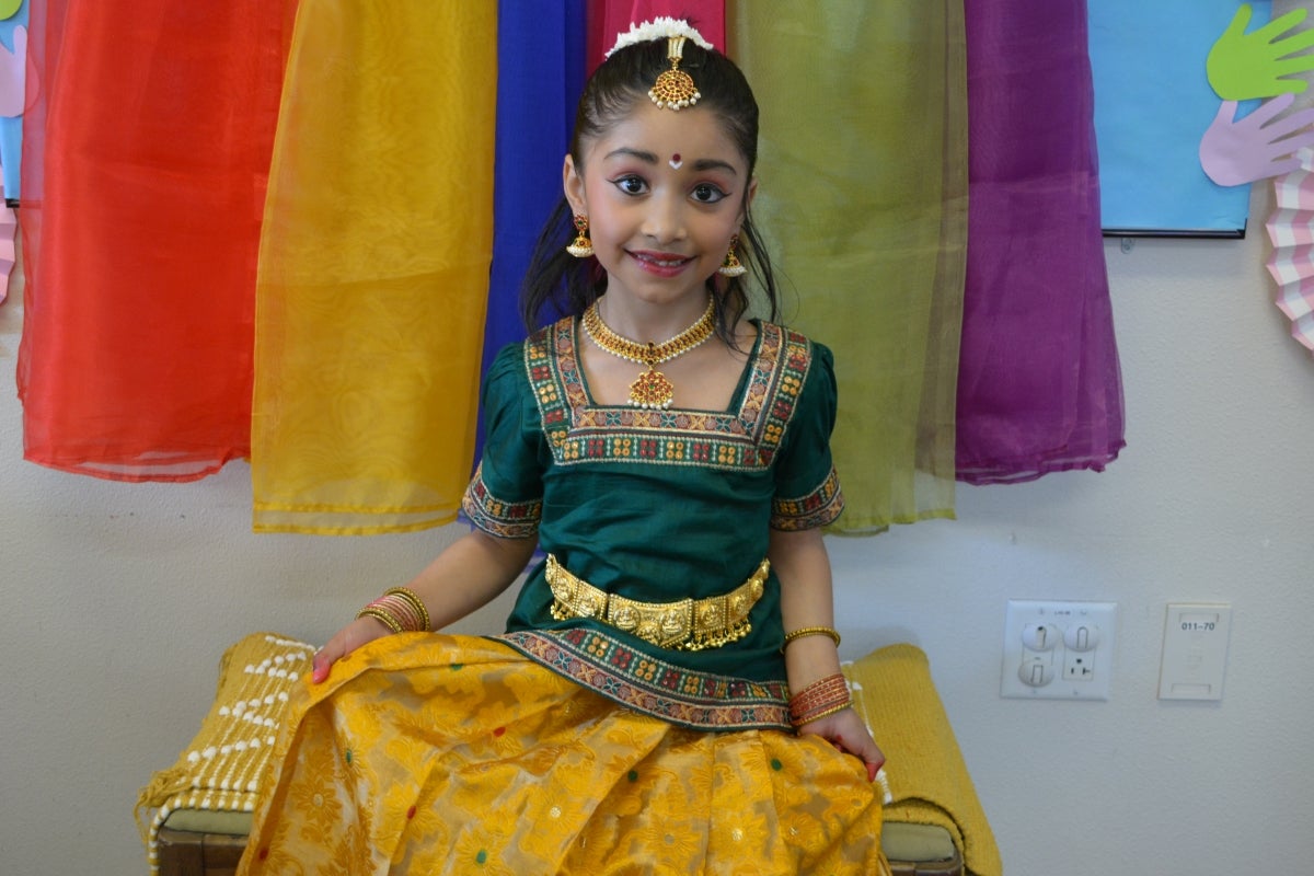 A young girl in traditional South Indian attire, sitting in front of colorful sheer fabric.