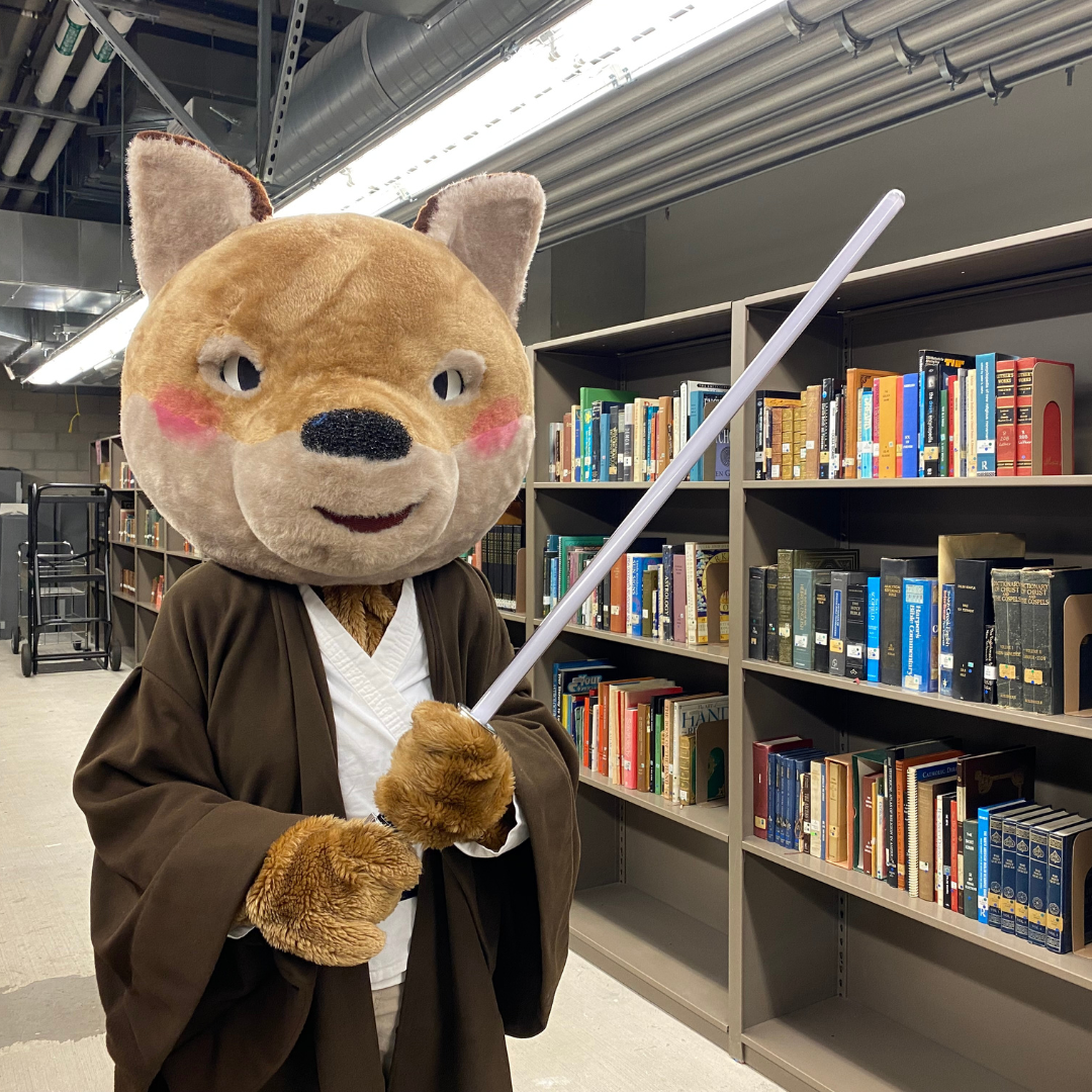 Photo of library mascot ODI cosplaying in a jedi robe holding a lightsaber in front of a row of book shelves