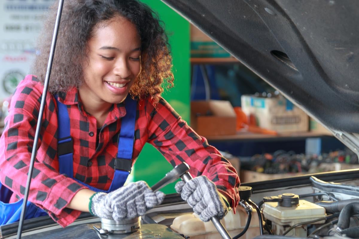 Young adult wearing coveralls and gloves working in car engine bay with tools in hand