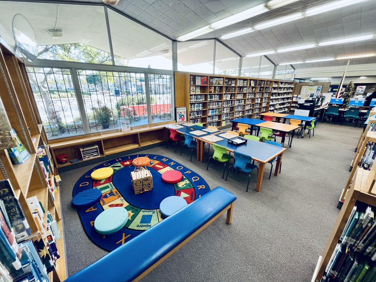 inside the library, the Children's Area Seating and Play Nook sit up against tall windows and wall shelving. The Play Nook has a blue bench beside an oval blue rug with the alphabet and colorful images. On top of the rug sits round colorful play mats and a play block in the center. The rest of the Children's Area has shelving units and 6 children's sized tables and chairs with puzzles and coloring pages sitting on top.
