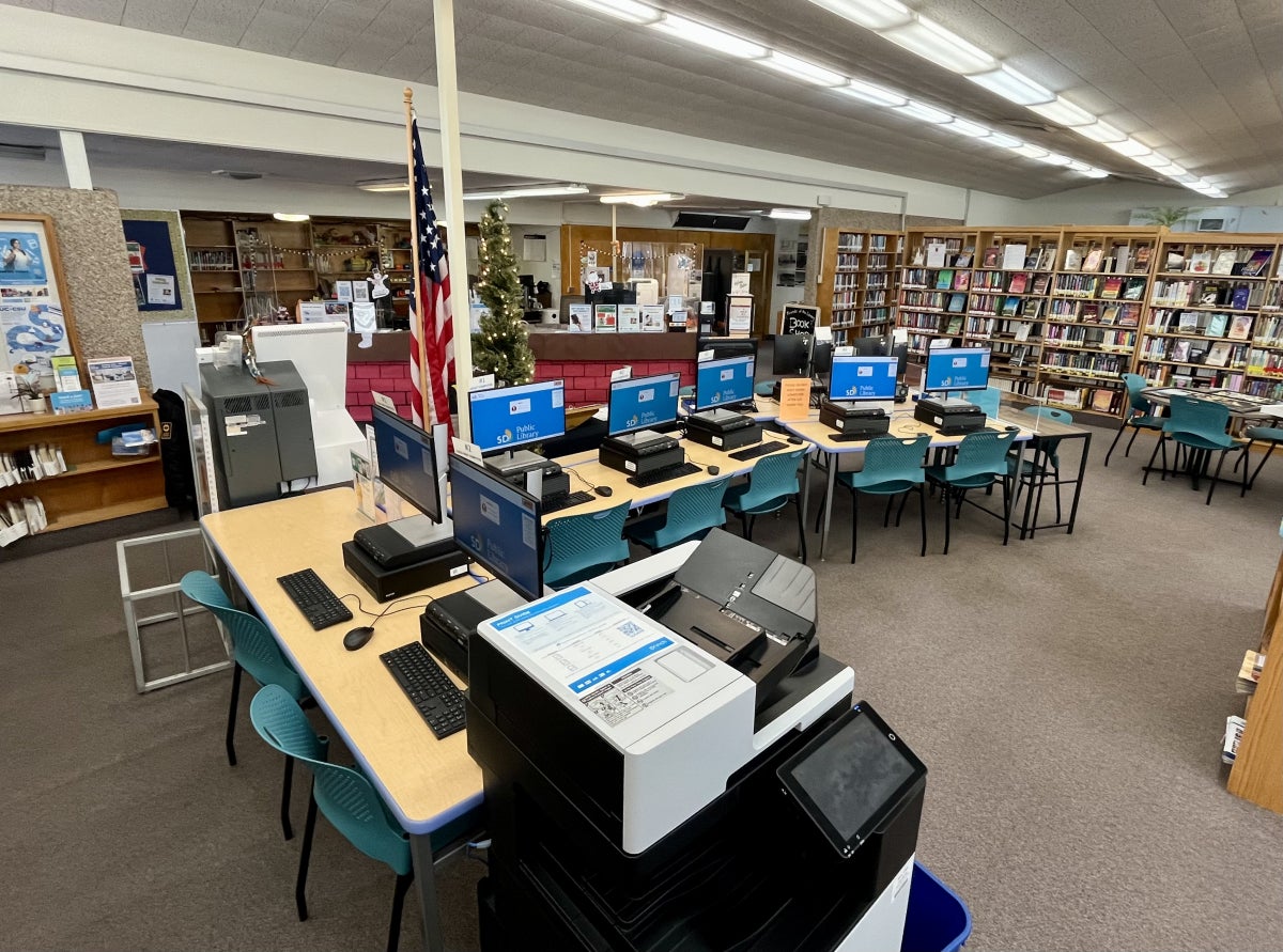 Rows of public library computer stations for youth and adults with teal chairs are surrounded by bookshelves, an American flag, and a front service desk. A large printer/copier machine sits at the end of the workstations.