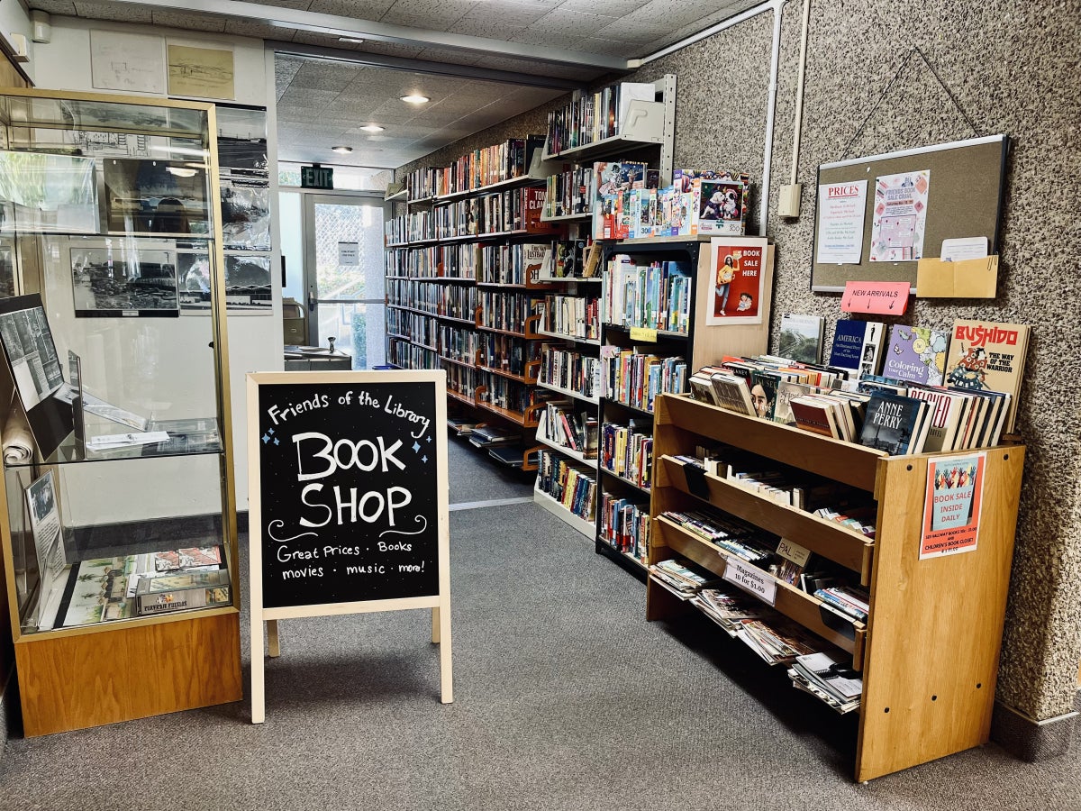 an A frame folding chalkboard sign has the words "Friends of the Library Book Shop. Great prizes, Books, Movies, Music, More!" Beside the sign is a display case full or branch historical photographs and papers. Behind the sign is a short hallway with books on walled shelves and a unit with drawers that pull out, also filled with books, DVDs, and Magazines—all for sale. 