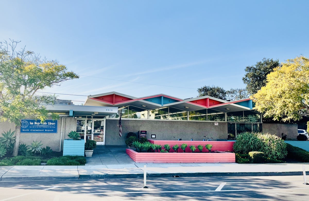 street view of the North Clairemont Library on Clairemont Drive. Tall trees on either side with various succulents and bushes in front. The building is a single story and features mid-century modern Googie architecture design with teal and salmon pink coloring. The roof features bold triangular shapes.