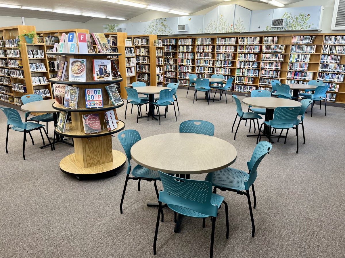  6 large round tables with 4 teal chairs at each table spread out in an open space surrounded by shelves of books along the walls and books stacks. A tall book display of Staff Picks features cookbooks.