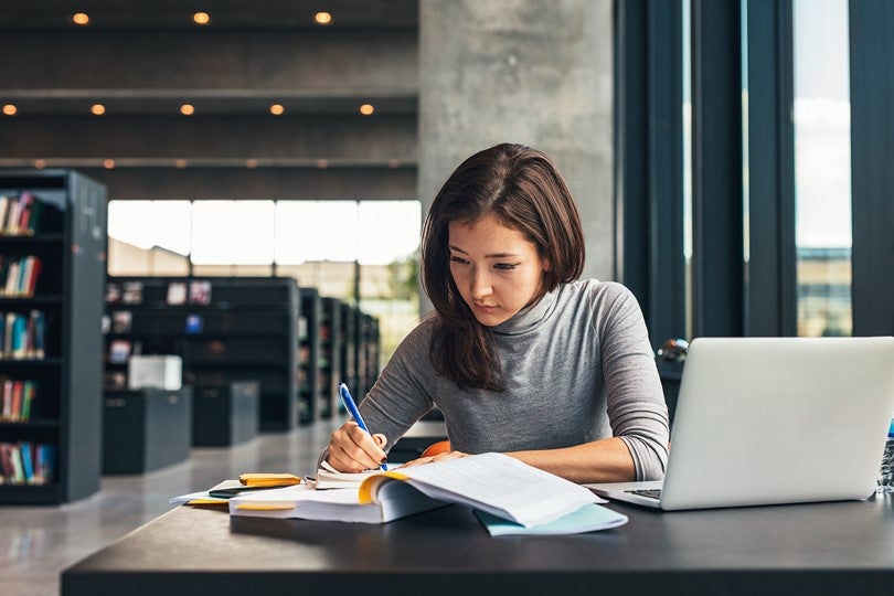 long haired woman seated at desk writing something with book and laptop open
