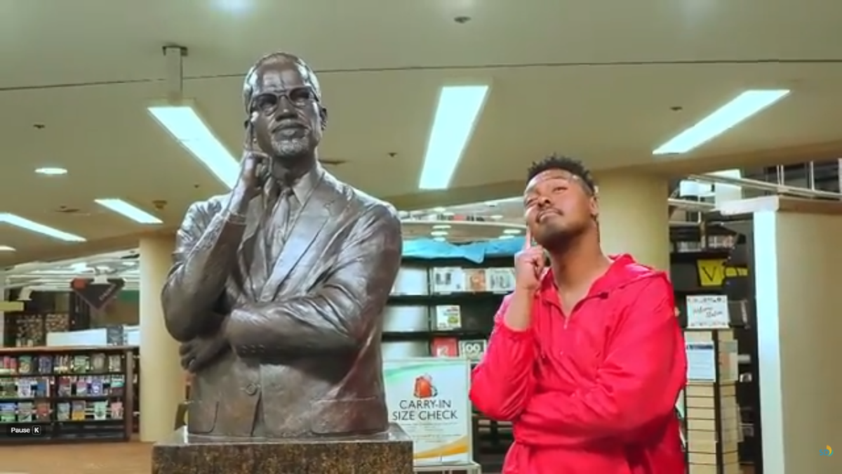 a young man imitates the contemplative pose of a bronze Malcolm X bust inside the library
