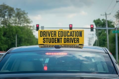 Large yellow sign attached to top of car stopped at red light stating “driver education student driver” in large text.