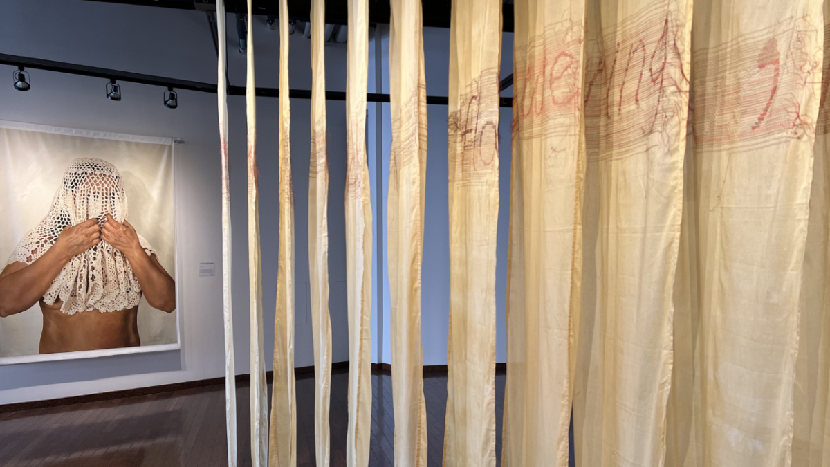 Closeup artworks in the Mother Tongue exhibition. The word “flowering” is seen embroidered across multiple draped panels. In the background is a large-scale painting by Leila Khalizadeh Aghdami of a nude woman wearing a white crocheted veil and clutching it with her hands.