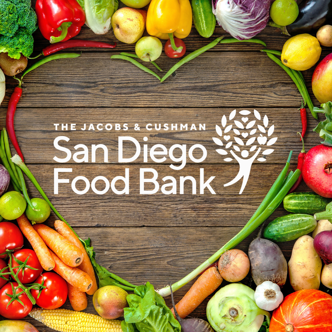 Colorful vegetables are arranged on a brown wooden table in the shape of a heart. San Diego Food Bank Logo is in the middle that is comprised of a white figure reaching up to a tree. The text reads as The Jacobs & Cushman San Diego Food Bank.