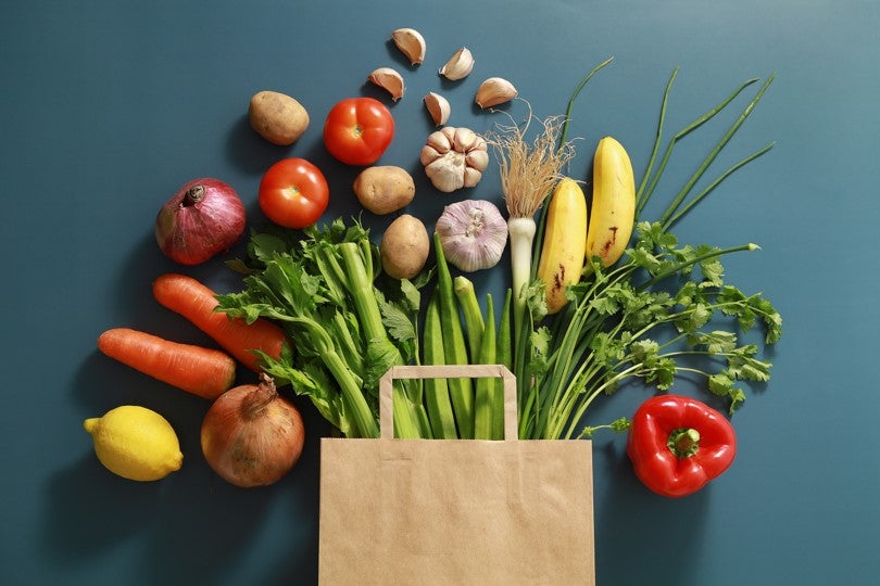 Various vegetables surrounding a brown grocery bag on a blue background.
