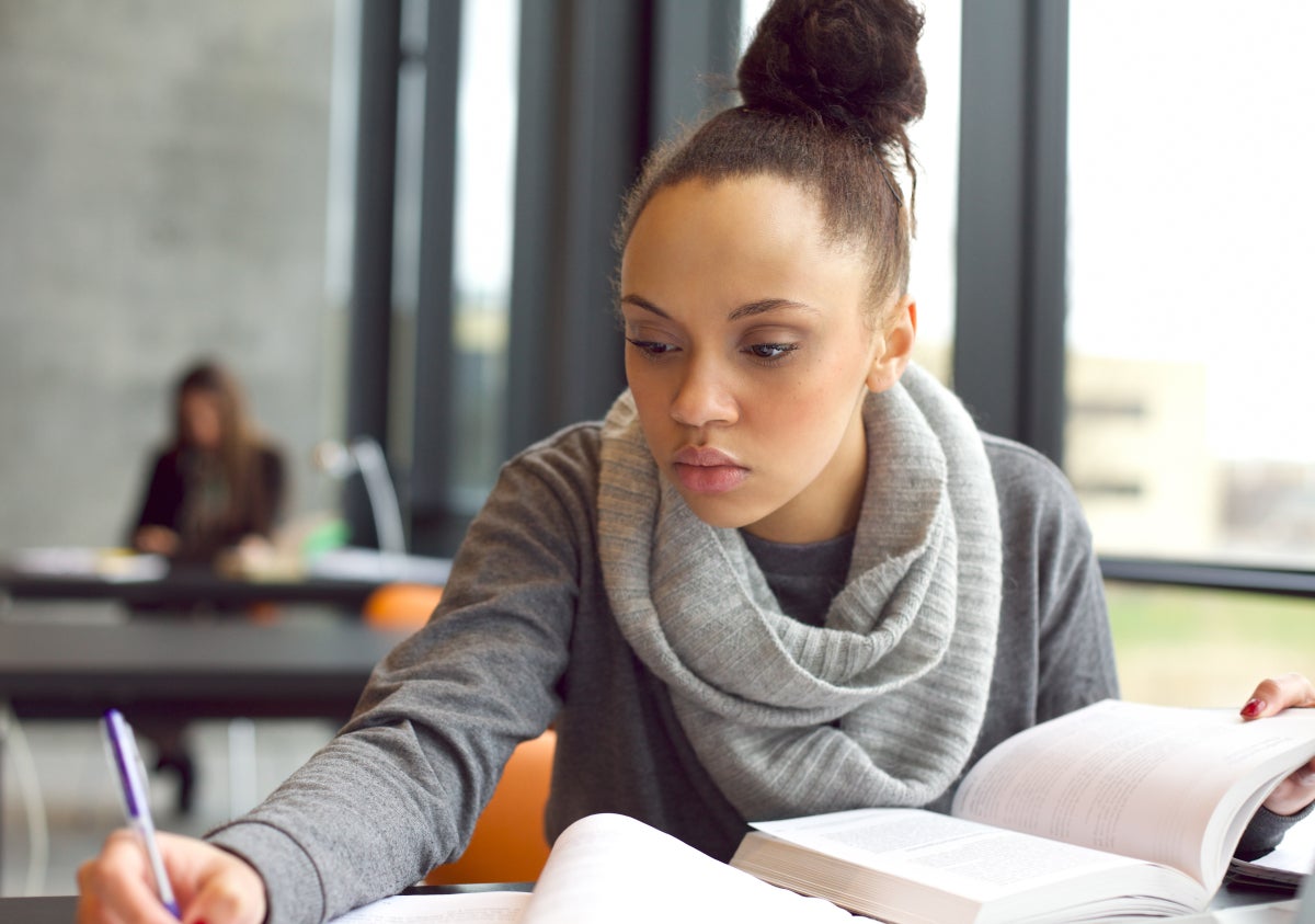 Woman holding a book open while sitting and writing with a pen.