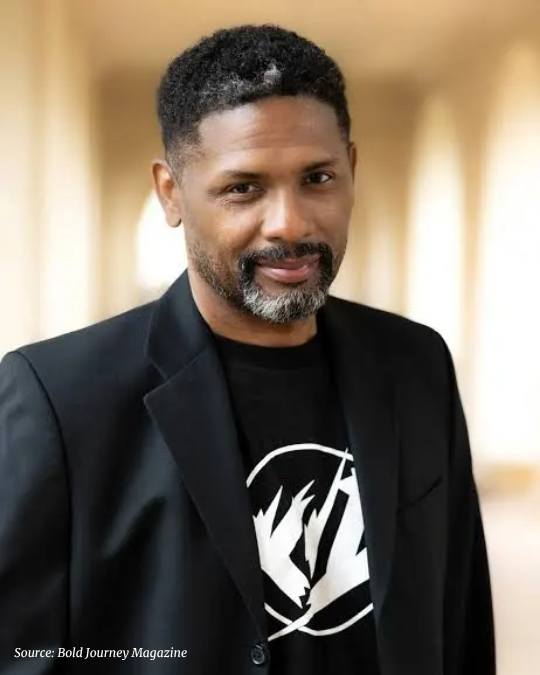 Photo portrait of an African American man with a neat beard wearing a blazer and tshirt with a colonnade backdrop.  