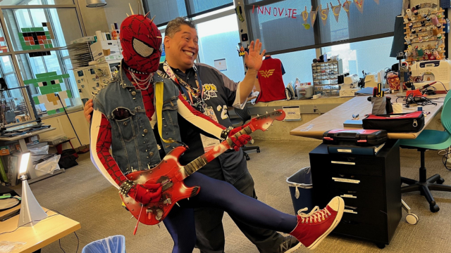 Spiderman in a punk outfit wears denim, spikes on his head and red converses while playing a spiderman-styled guitar.