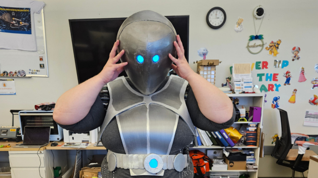 A woman in a robot cosplay holds up her metal helmet lit up with electric blue eyes.