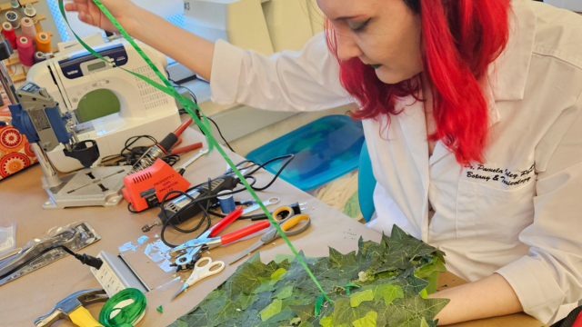 A red-haired woman threads a ribbon into the leaf-covered bodice of her Poison Ivy costume.