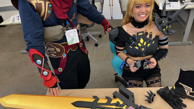 A  petite Asian woman in armor and a wig smiles at the camera while holding accessories for her costume. A giant foam sword lies on the table alongside gloves and cuffs.