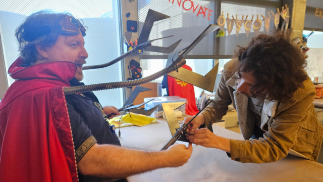 A  middle aged man in a cape sits at a table so the machine claws of his costume can be worked on by an IDEA Lab staff member. 