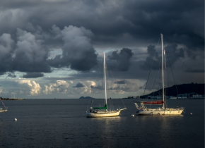 Two sailboats at sea illuminated at dusk on calm water beneath dramatic dark storm clouds.