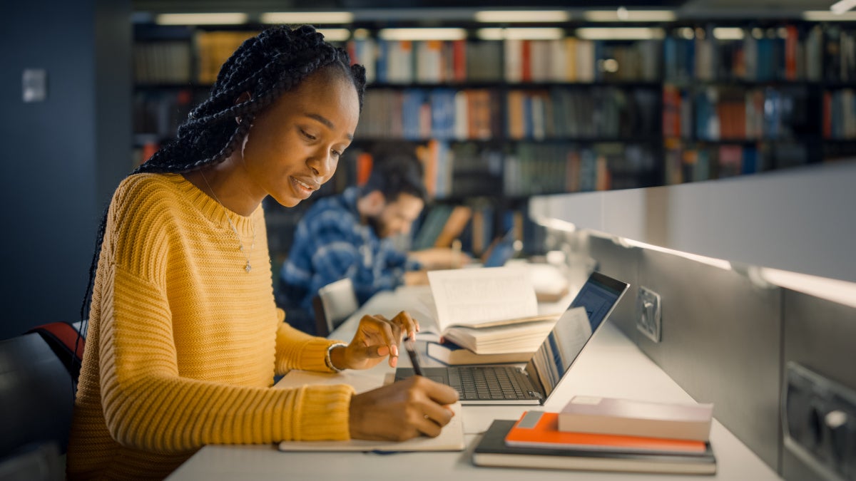 a student in a yellow sweater is studying in a library on her laptop.
