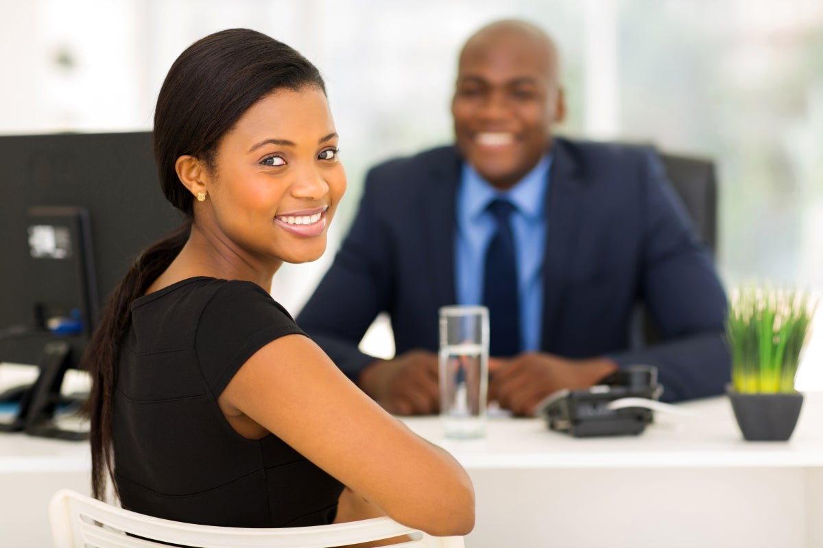 A woman in a black dress is sitting at a desk having a meeting with a man in a blue suit in an office. 