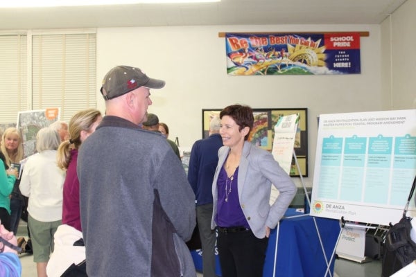 Person wearing a hat looking at a posterboard for De Anza Revitalization Plan. A person wearing a blazer is smiling.
