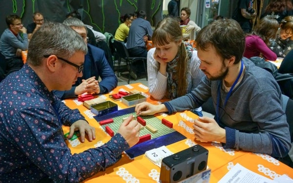 Four people at the table looking at red and tan blocks on a grid. The room is filled with other people.