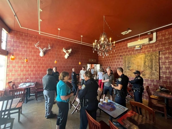 San Diego police officers speaking to community members inside of a cafe. The cafe walls are patterned and red and a chandelier hangs above the group.