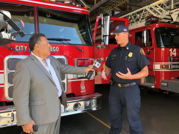 A journalist holds a microphone up to a firefighter. Both people are standing in front of a fire apparatus.