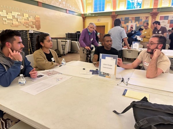 Four pople at a table with notes. The table has a "Urban Design Implementation" label. Many people are in the background having discussions at other tables.